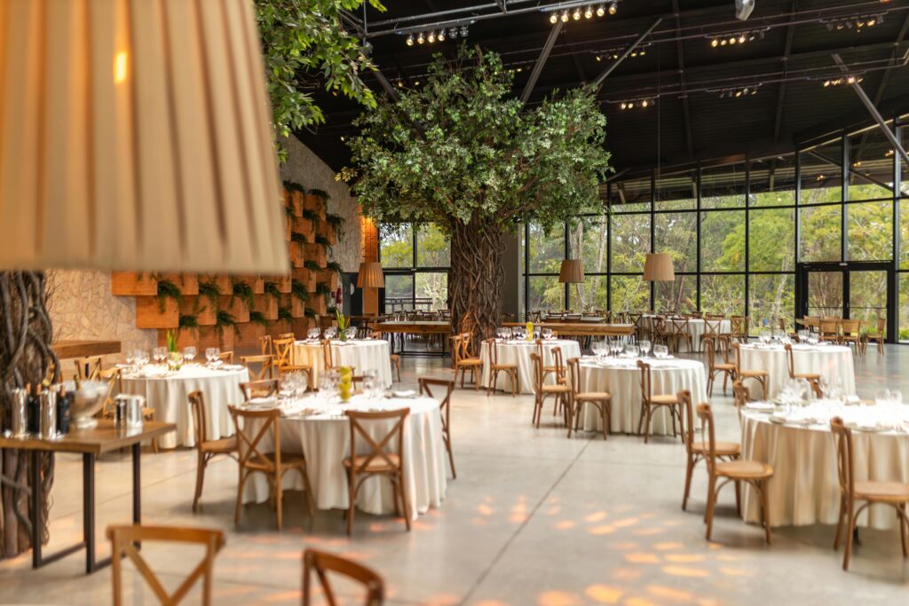 pexels-photo-17057017-17057017 Spacious event hall featuring round tables, chairs, and indoor trees. Ideal for weddings or conferences.